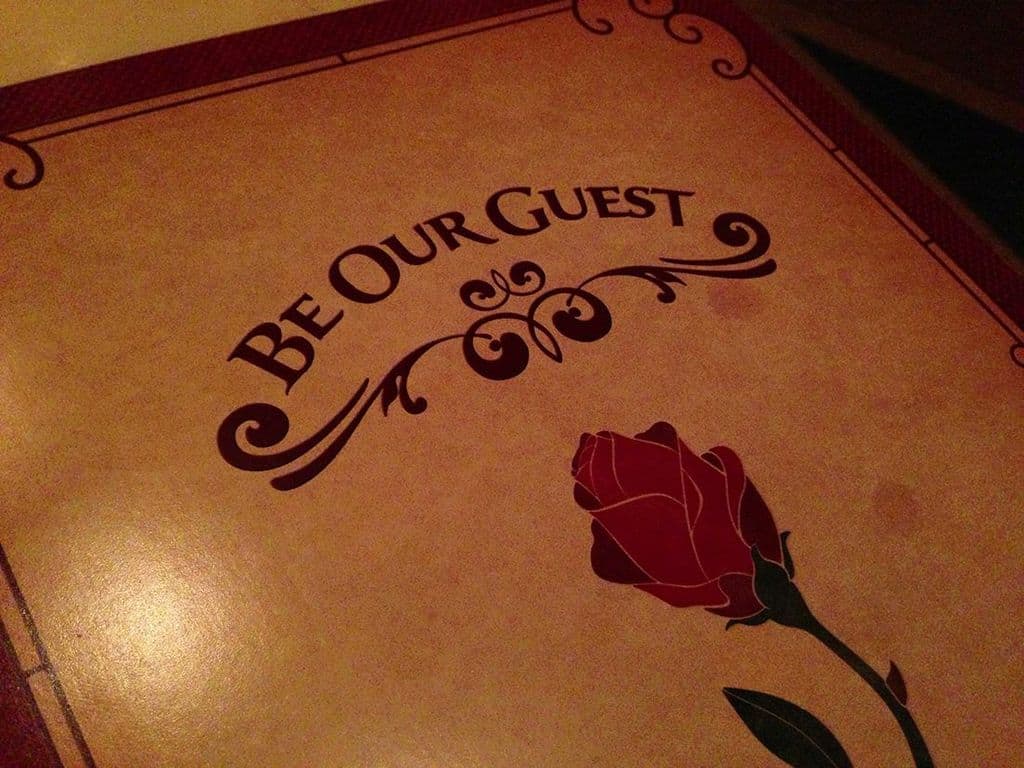 Be Our Guest" menu cover with a red rose on tan, evoking the magic of Disney dining for theme park fans and planners.