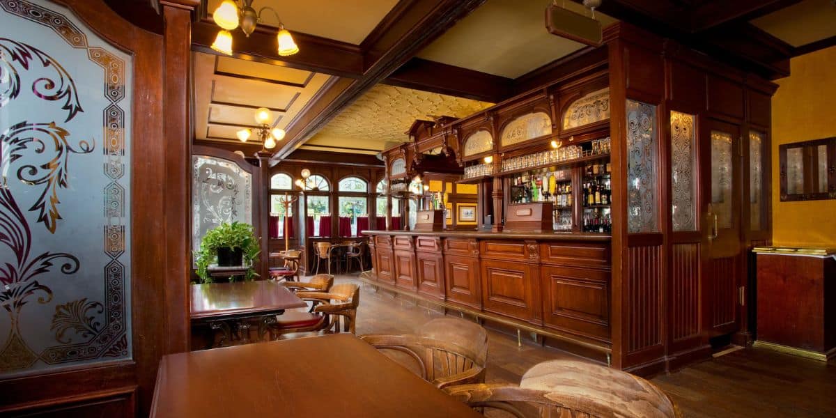 A cozy pub interior with wooden furniture and warm lighting. There is an ornate wooden bar with stools, and tables with chairs in the foreground. Decorative glass panels and hanging lights add to the inviting atmosphere.