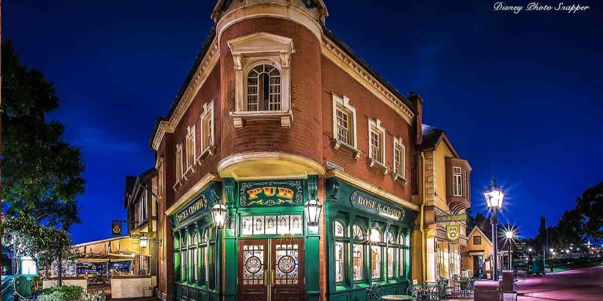 A charming, illuminated pub on a street corner at night. The building features red brick with green and wood accents, ornate windows, and signage. Street lamps and surrounding trees frame the scene under a deep blue sky.