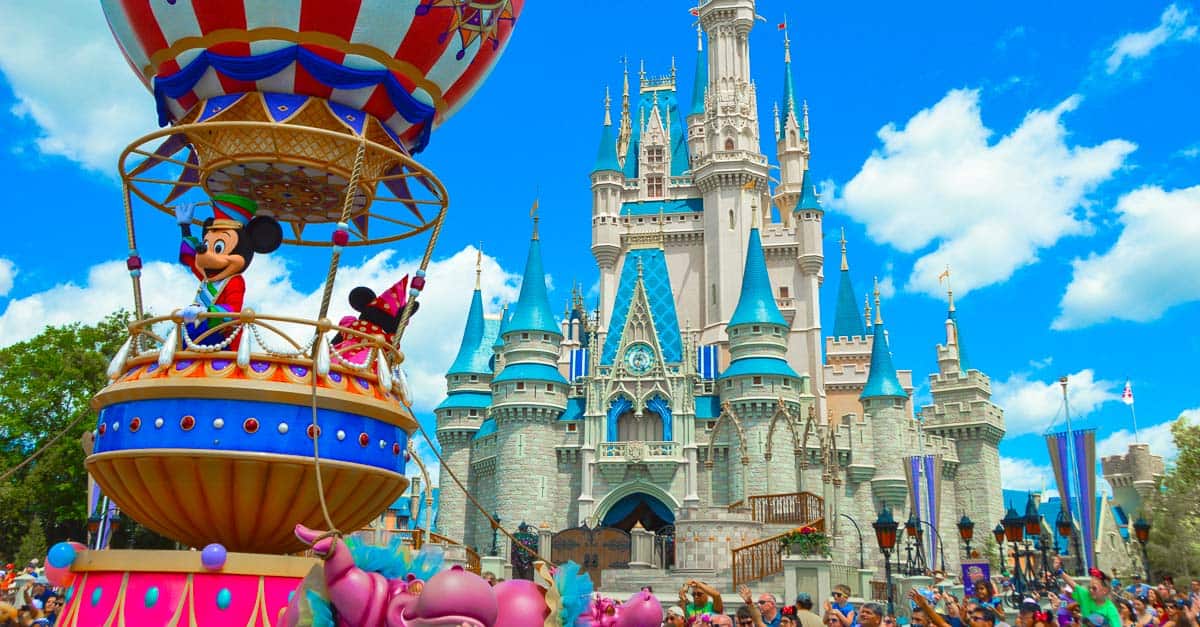 A colorful parade float with cartoon characters in front of a large, ornate castle at a theme park, surrounded by a crowd under a bright blue sky with some clouds.
