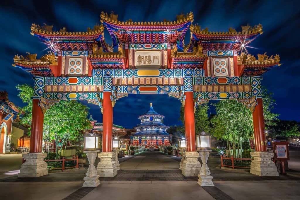 Epcot’s China Pavilion: vibrant, illuminated Chinese archway at night, bamboo-lined path, temple and glowing lights behind.