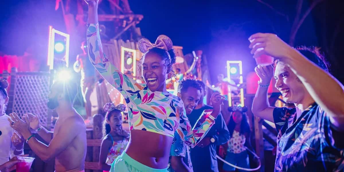 A group of people dance and celebrate at a vibrant, colorful outdoor party at night, with bright lights and festive decorations in the background. A smiling woman in a colorful outfit is in the center, raising her arms in joy.