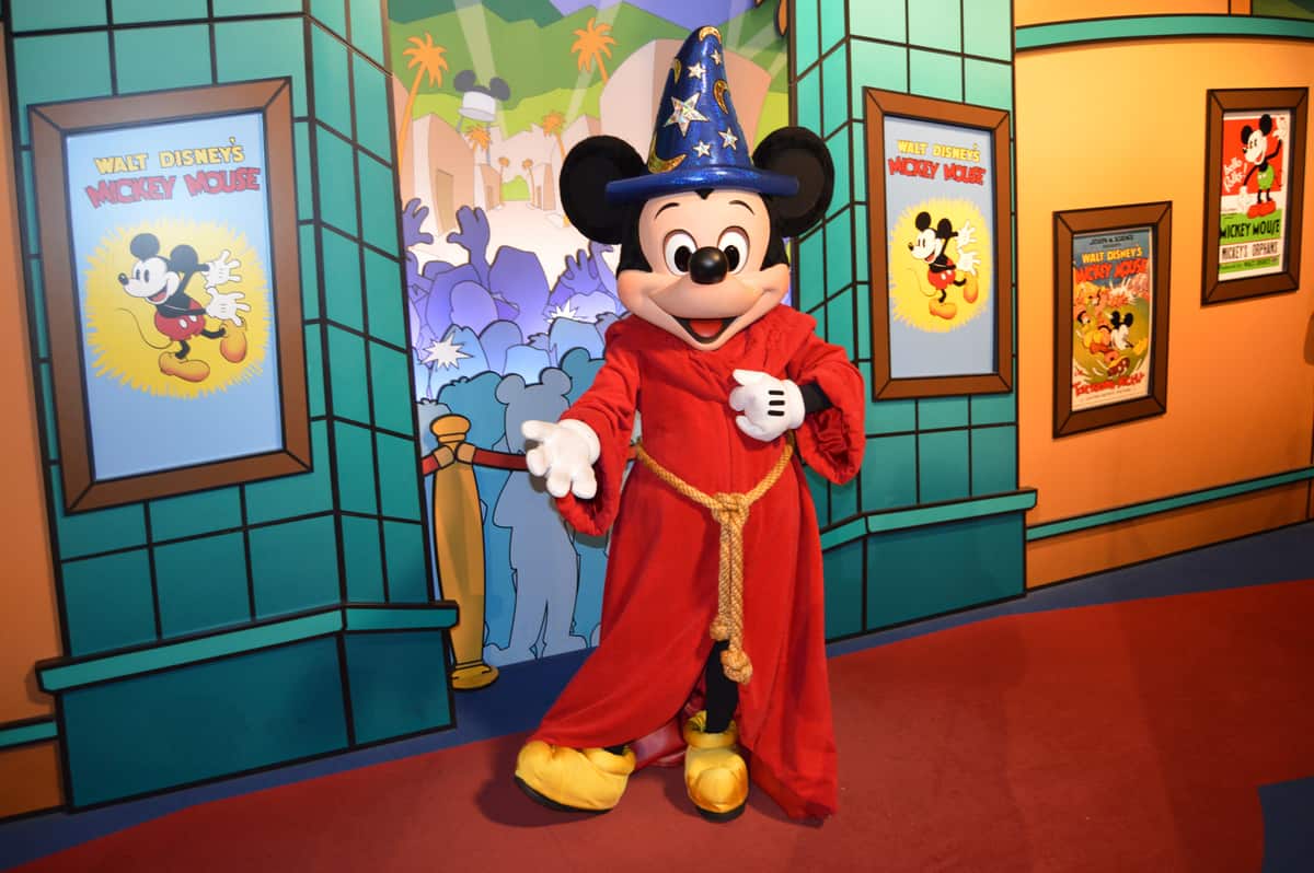 Mickey Mouse in his red robe and blue wizard hat greets guests with magical energy amid vibrant Disney World backdrops.