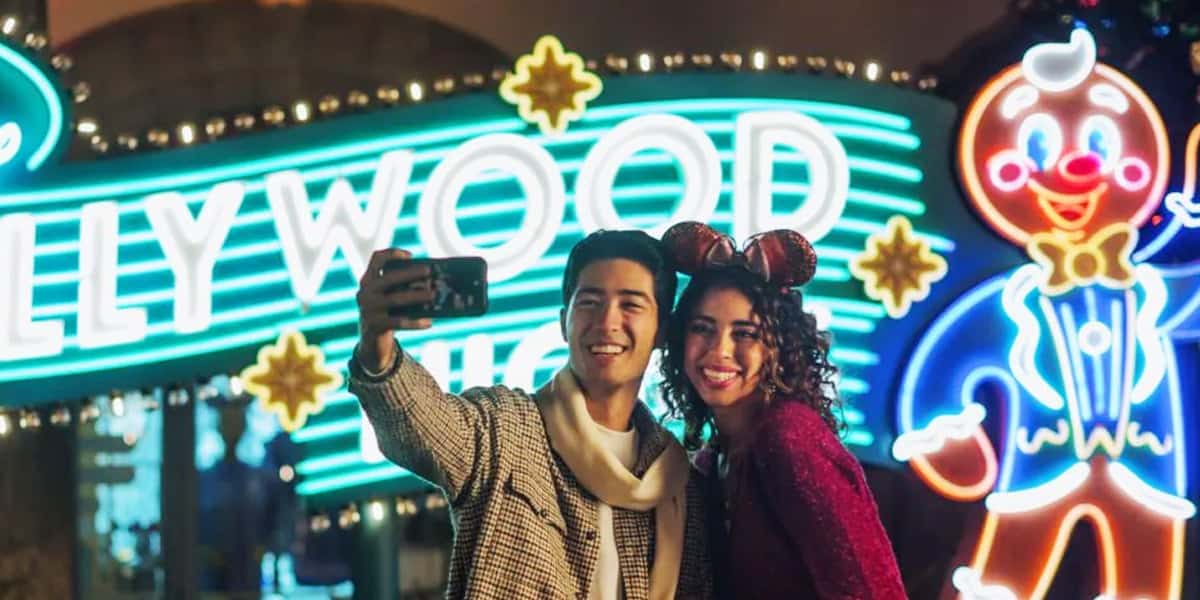 A smiling young couple in festive outfits takes a selfie at night in front of a brightly lit Hollywood sign and a large glowing gingerbread man decoration.