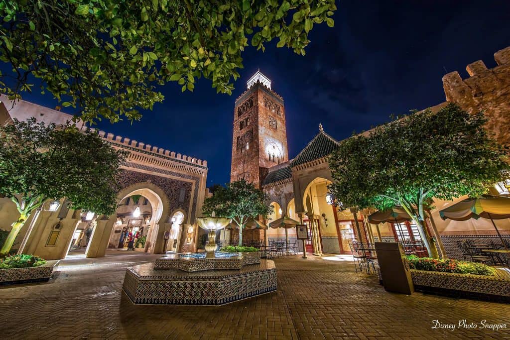 Magical nighttime scene at EPCOT’s Morocco Pavilion with a sparkling fountain, lush trees, grand arches, and the Koutoubia tower.