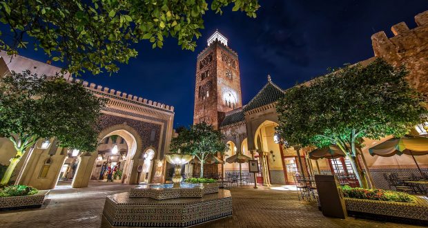 Magical nighttime scene at EPCOT’s Morocco Pavilion with a sparkling fountain, lush trees, grand arches, and the Koutoubia tower.