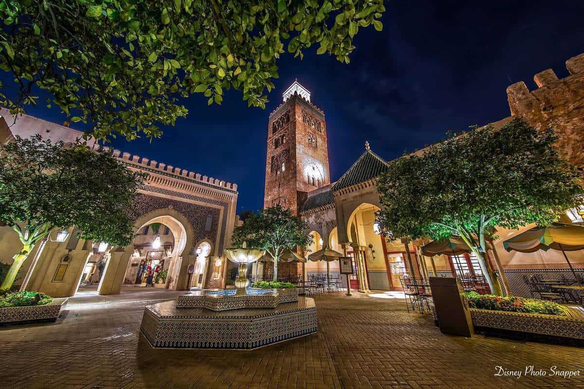 Magical nighttime scene at EPCOT’s Morocco Pavilion with a sparkling fountain, lush trees, grand arches, and the Koutoubia tower.