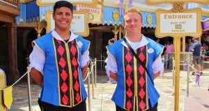 Disney World Cast Members in blue vests stand at a ride entrance, holding a sign showing a 150-minute wait time.