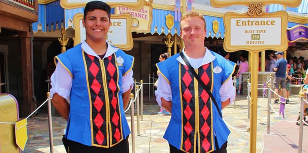 Disney World Cast Members in blue vests stand at a ride entrance, holding a sign showing a 150-minute wait time.
