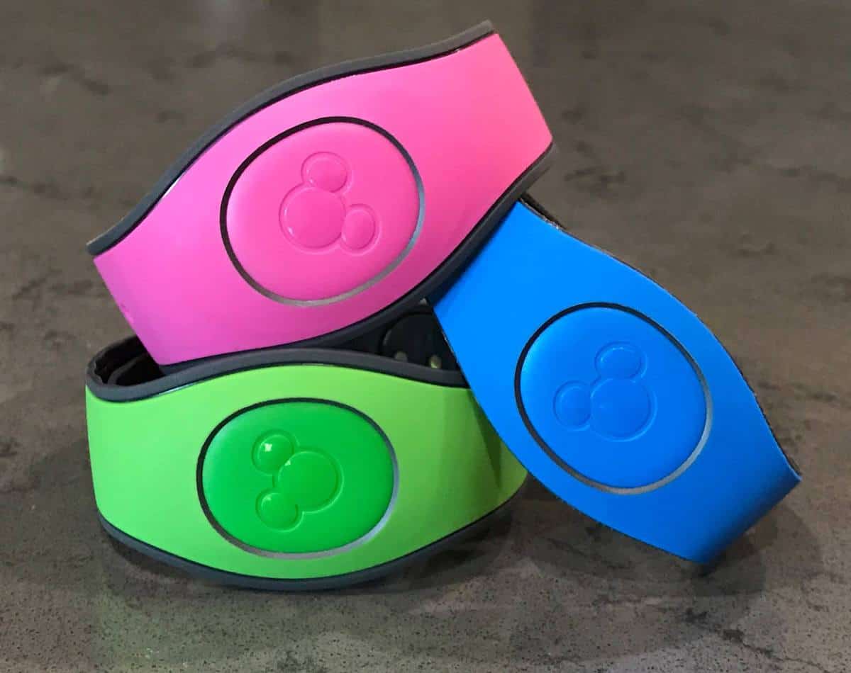 Pink, green, and blue Disney MagicBands stacked on gray, highlighting how park guests can personalize bands with themed accessories.