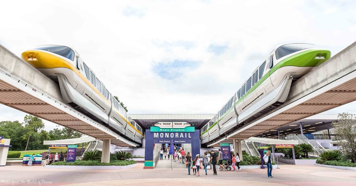 Disney World monorails glide above guests entering Magic Kingdom, passing under the iconic "Magic Kingdom Monorail" sign.
