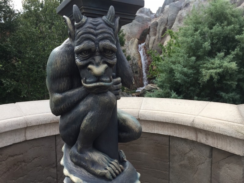 A gargoyle clings to a column amid lush greenery and a waterfall, channeling the enchanted vibes of iconic Disney venues.