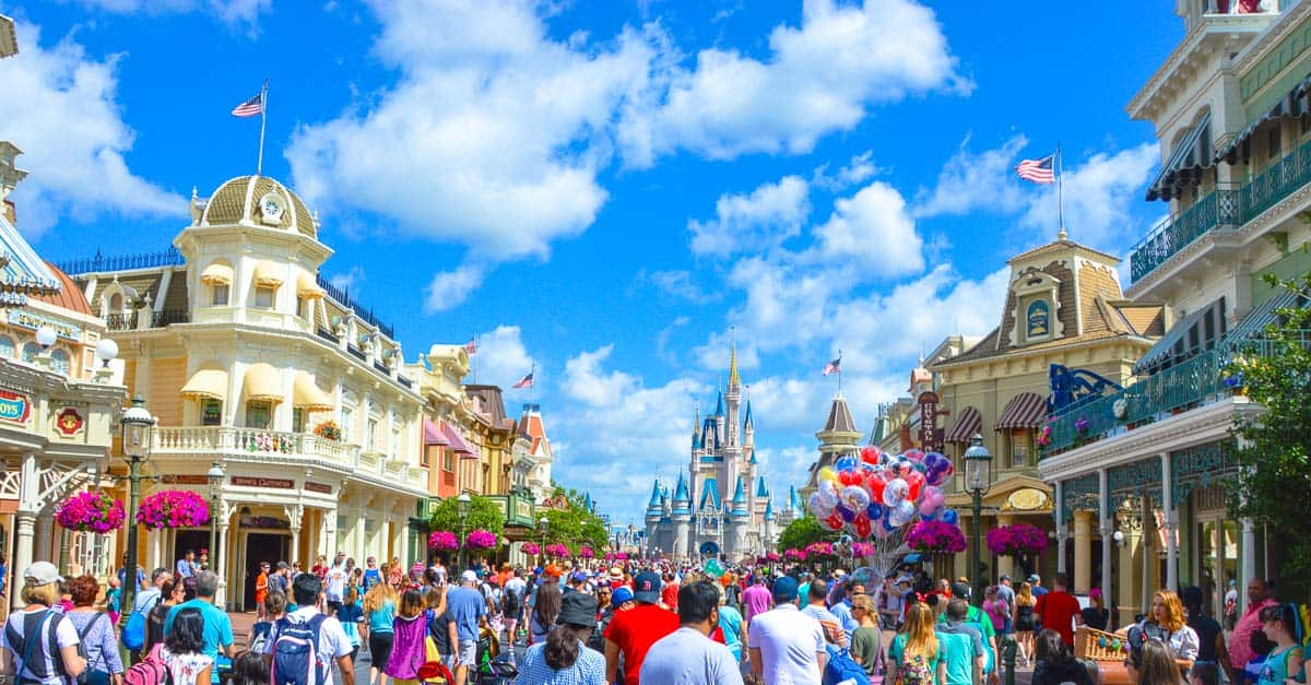 Guests stroll down Main Street, U.S.A. toward Cinderella Castle at Disney World under a vibrant sky with puffy white clouds.