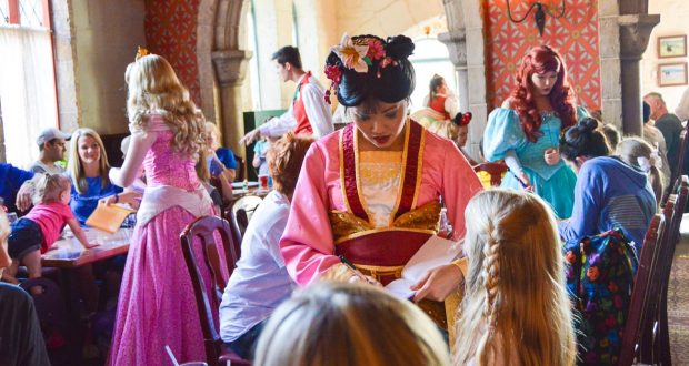 In a lively Disney dining hall, Aurora in her pink gown greets a young girl with braids during a magical princess meet-and-greet.