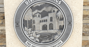 Disney Springs plaque with "The Source of Inspiration" motto, scenic building and lush plants—perfect for Disney World insiders.