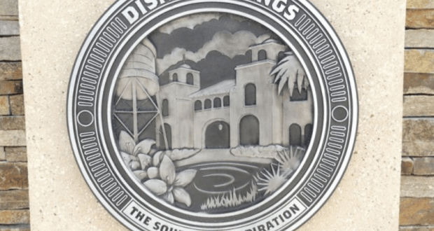 Disney Springs plaque with "The Source of Inspiration" motto, scenic building and lush plants—perfect for Disney World insiders.