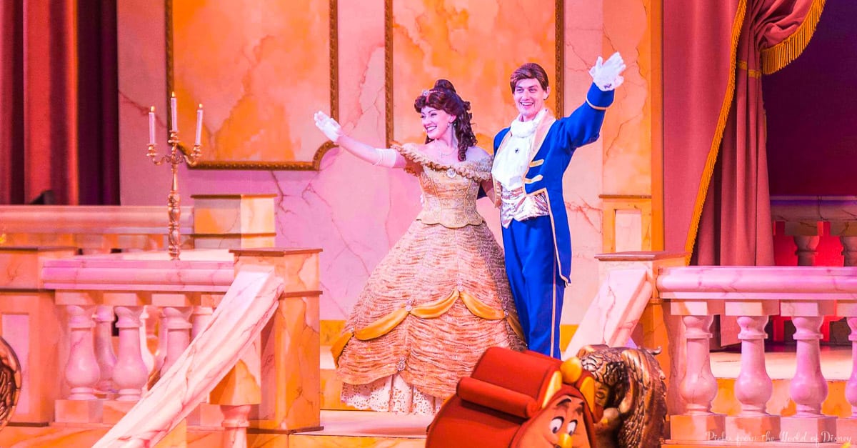 Two performers dressed as Belle in a gold gown and the Beast in a blue suit stand on stage, smiling and waving, with elegant castle-like decor and warm lighting in the background.