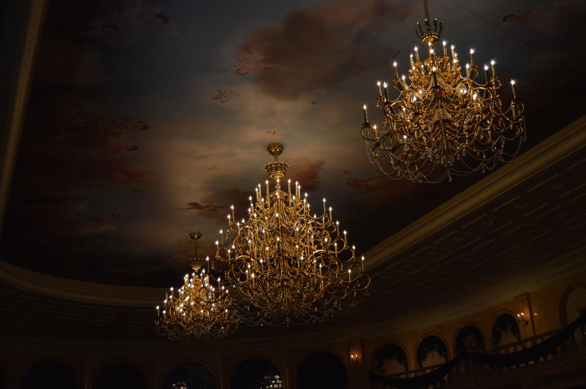 Three ornate chandeliers glow beneath a dreamy, cloud-painted ceiling, evoking the enchantment of Disney World dining magic.