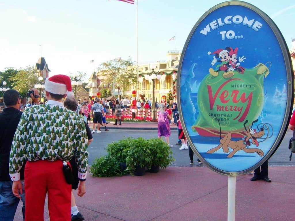 Festive guest poses by Mickey's Very Merry Christmas Party sign at Disney World entrance, kicking off 2025 holiday celebrations.