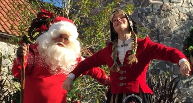 Santa and a Disney Cast Member in holiday costumes pose and smile by a stone building, celebrating festive magic at Disney Parks.