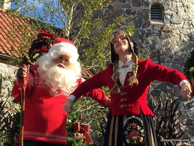 Santa and a Disney Cast Member in holiday costumes pose and smile by a stone building, celebrating festive magic at Disney Parks.