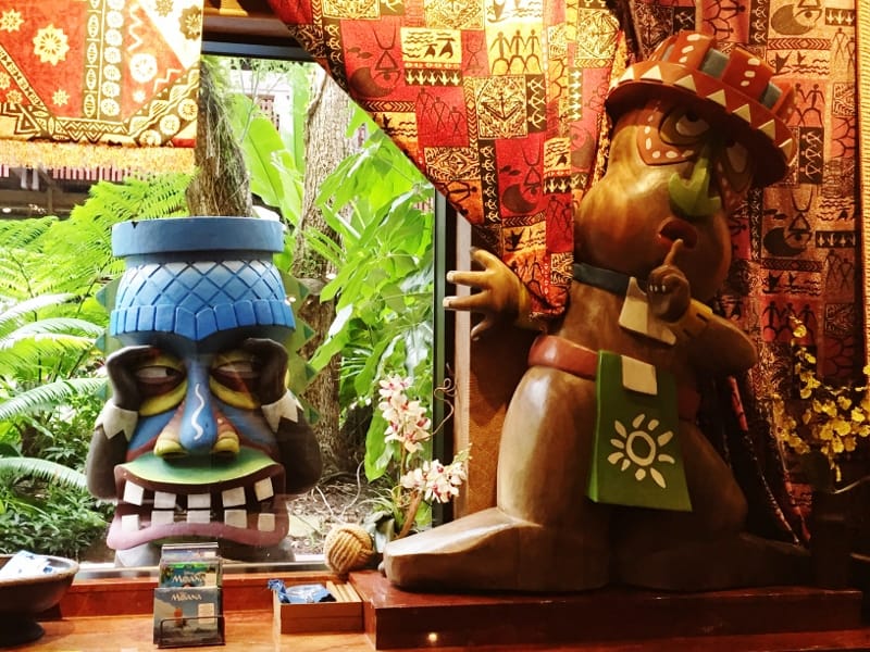 Vibrant tiki statues channel Disney Resort vibes, framed by lush tropical plants and bold fabrics like at Polynesian Village.