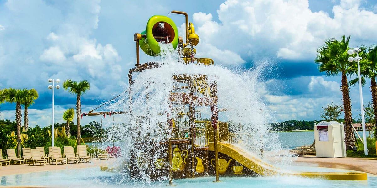 A large water play structure with slides and a giant bucket dumping water in a splash zone, surrounded by palm trees, lounge chairs, and a scenic lake under a partly cloudy sky.