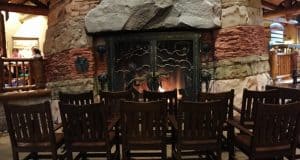 Six wooden rocking chairs circle a glowing stone fireplace, capturing the lodge-style warmth found at Disney Resort hotels.