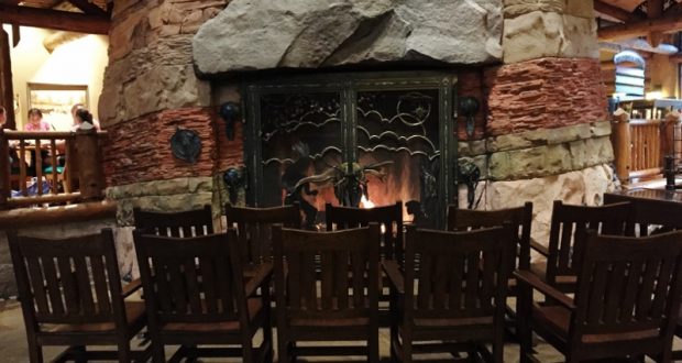 Six wooden rocking chairs circle a glowing stone fireplace, capturing the lodge-style warmth found at Disney Resort hotels.