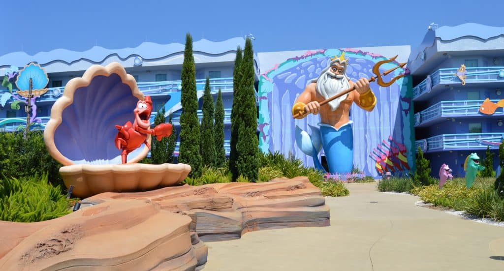 A colorful outdoor scene at a Disney resort featuring a large statue of Sebastian the crab in a seashell and a mural of King Triton with his trident, surrounded by greenery and whimsical, ocean-themed architecture.