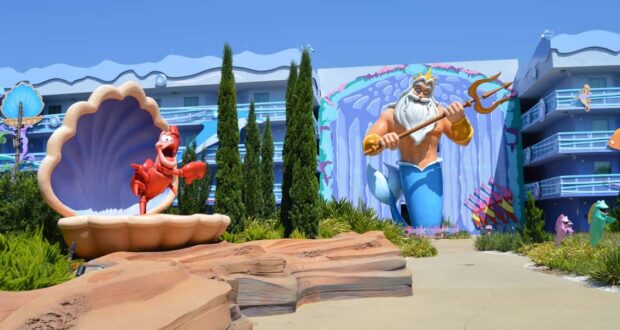 A colorful outdoor scene at a Disney resort featuring a large statue of Sebastian the crab in a seashell and a mural of King Triton with his trident, surrounded by greenery and whimsical, ocean-themed architecture.