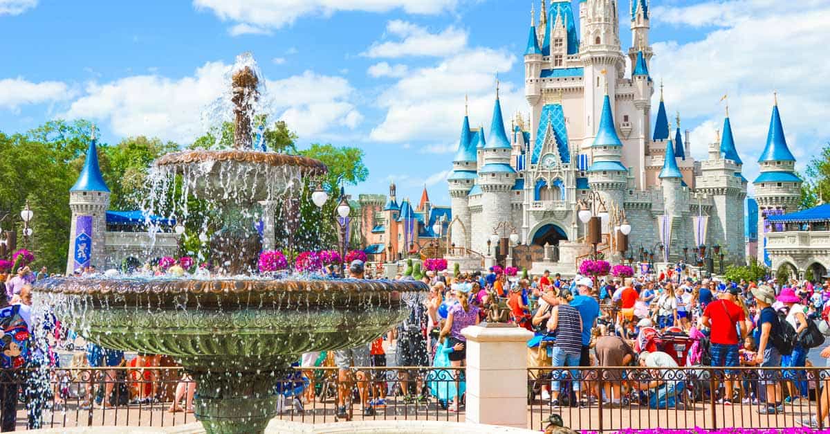 Guests gather by Cinderella Castle with its iconic blue spires and sparkling fountain on a sunny day at Disney World.