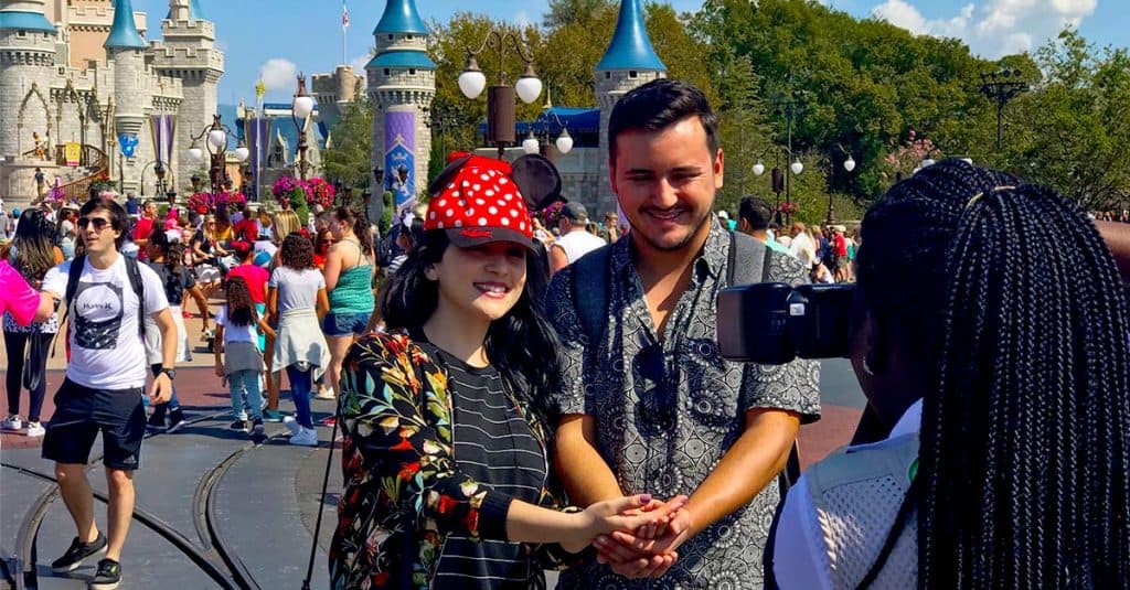 Guests smile for a Memory Maker photo in front of Cinderella Castle at Disney World, surrounded by lively crowds and a pro photographer.