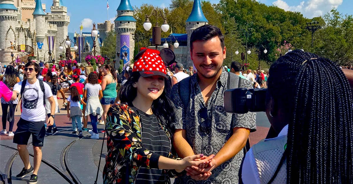 Guests smile for a Memory Maker photo in front of Cinderella Castle at Disney World, surrounded by lively crowds and a pro photographer.