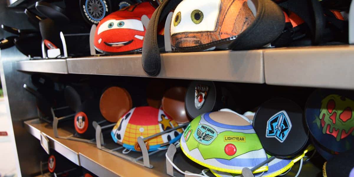 Colorful Disney-themed Mickey Mouse ear hats, including designs inspired by Cars and Toy Story, are displayed on metal shelves in a store.