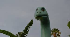 Gertie the Dinosaur munches leaves by Echo Lake, framed by palm trees and dramatic clouds at Disney’s Hollywood Studios.