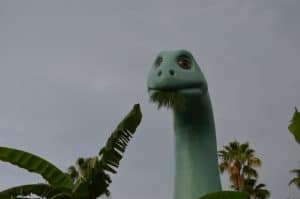 Gertie the Dinosaur munches leaves by Echo Lake, framed by palm trees and dramatic clouds at Disney’s Hollywood Studios.