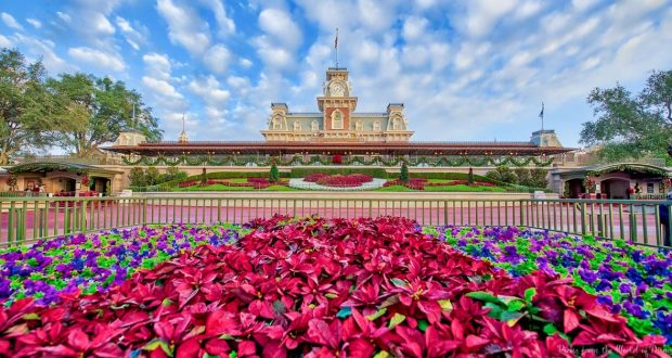 Vibrant flowerbeds welcome guests to the majestic Main Street U.S.A. train station and clock tower beneath a bright Disney sky.