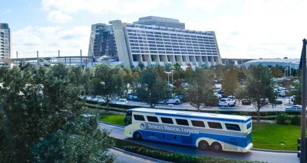 A blue and white "Disney's Magical Express" bus drives on a curved road in front of the large, modern Disney’s Contemporary Resort hotel with many parked cars and trees surrounding the area.