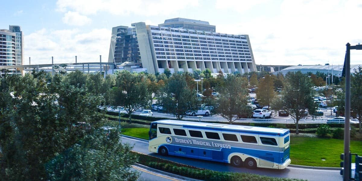 A blue and white "Disney's Magical Express" bus drives on a curved road in front of the large, modern Disney’s Contemporary Resort hotel with many parked cars and trees surrounding the area.
