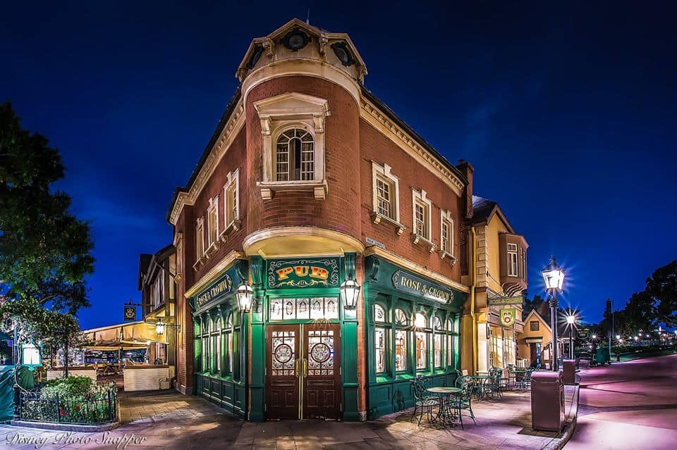 A lively, warmly lit British pub on a Disney street corner at night, outdoor seating, deep blue sky—evoking Disney dining magic.