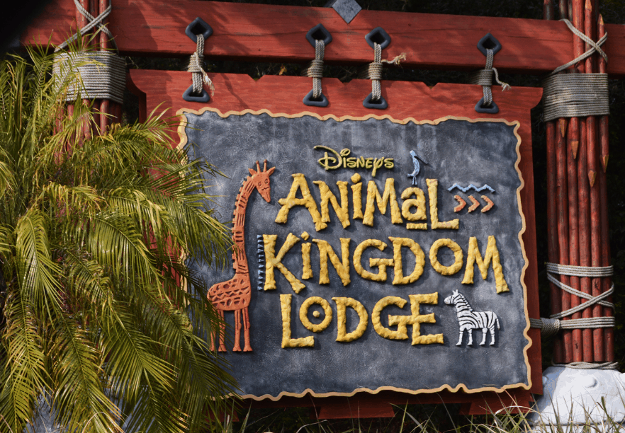 Disney’s Animal Kingdom Lodge sign with giraffe and zebra art, surrounded by tropical greenery near bustling Kidani Village.