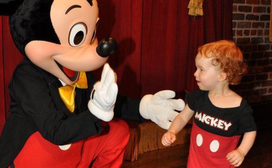 A person in a Mickey Mouse costume greets a smiling toddler wearing a Mickey Mouse-themed outfit. They are indoors with a red curtain and brick wall in the background.