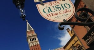 Tutto Gusto Wine Cellar sign and classic Italian facades at Epcot’s Italy Pavilion beneath a vibrant blue sky with fluffy clouds.