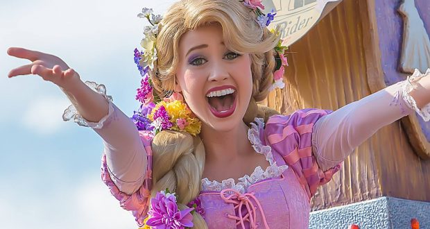 Rapunzel in her signature pink dress greets guests with a smile, posing by the "Wanted" Flynn Rider poster at Disney Parks.