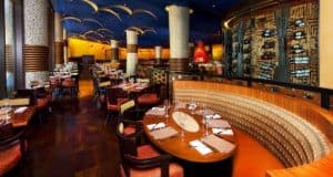 Upscale dining room with curved booths, set tables, golden lighting, and columns inspired by Jiko at Disney’s Animal Kingdom Lodge.