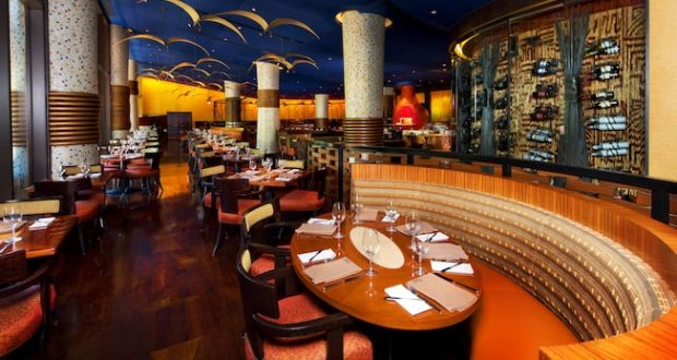 Upscale dining room with curved booths, set tables, golden lighting, and columns inspired by Jiko at Disney’s Animal Kingdom Lodge.
