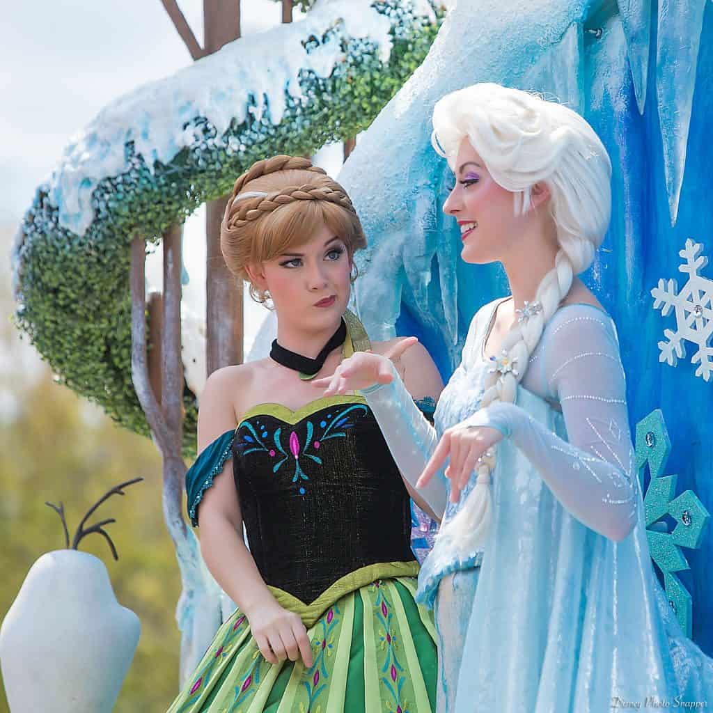 Anna and Elsa greet guests outside among snowy decorations and Olaf, ready to share fun Disney trivia at the park.