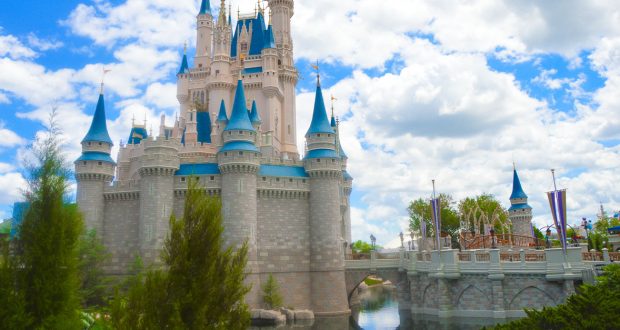 Cinderella Castle with blue turrets, surrounded by water and greenery beneath a partly cloudy sky at Walt Disney World.