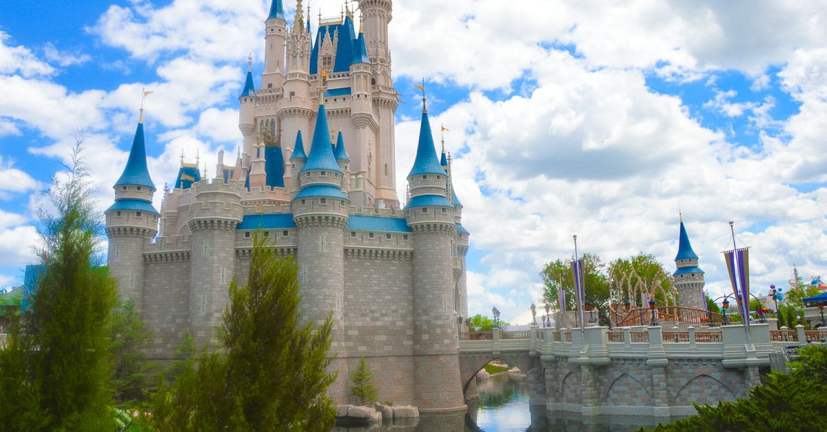 Cinderella Castle with blue turrets, surrounded by water and greenery beneath a partly cloudy sky at Walt Disney World.